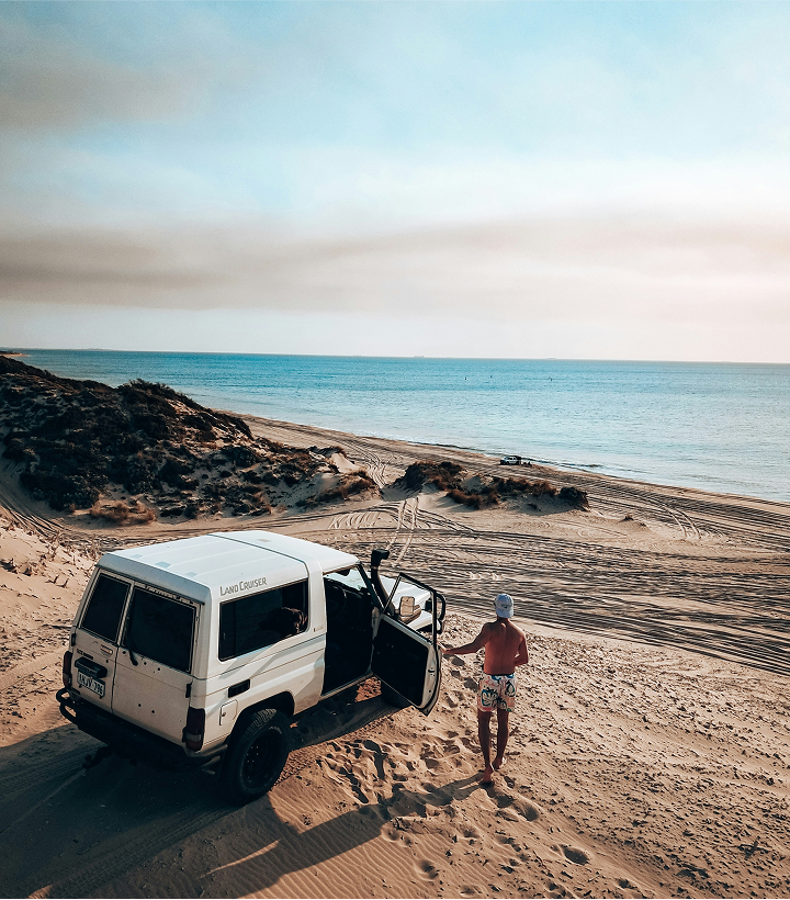 A white land cruiser parked on the beach with a door open. A man in his swim wear is standing next to the car looking over to the ocean.
