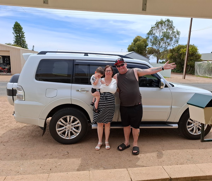 A happy looking family of three, mother, father and their son standing in front of a white SUV car.