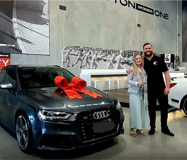 A brand new black Audio car is parked on the side with a red bow tie on top. A woman is standing next to a guy from Unrivalled Finance, smiling.